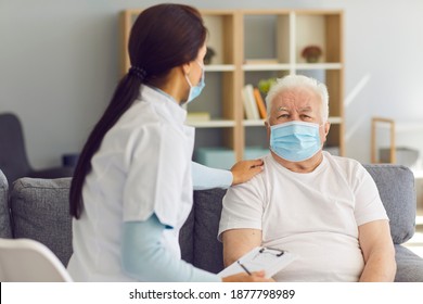 Seniors Are In Risk Group. Female Doctor Visiting Elder Male Patient At Home, Both Wearing Medical Face Masks. Healthcare Provider Supporting Old Man, Cheering Him Up During Coronavirus Pandemic