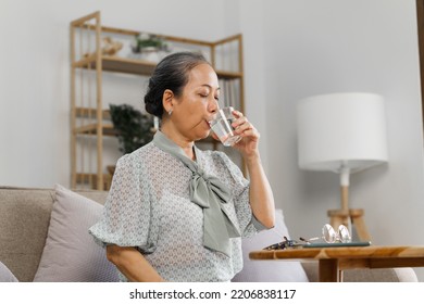 Senoir mature asian woman with glass of water taking pills, sitting on couch. - Powered by Shutterstock