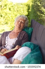 Senor Woman Sitting On Lounge Chair With A Book And Talking A Nap. Senior Lady Sleeping In Backyard With A Novel