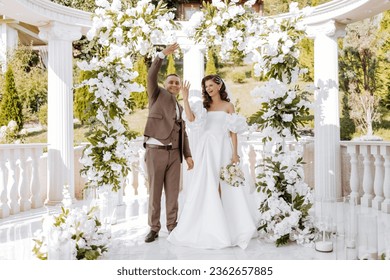 sensitive ceremony of the bride and groom. A happy newlywed couple stands against the background of a wedding arch decorated with fresh flowers. The bride and groom show off their wedding rings