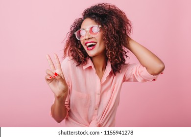 Sensual african lady in trendy vintage outfit enjoying good day on photoshoot. Fascinating lovable woman in sunglasses chilling in studio on pink background.