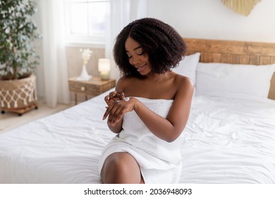 Sensual Afro woman sitting on bed in bath towel, applying hand cream, pampering her skin with moisturizing lotion at home. Pretty black lady making daily body care routines, indoors