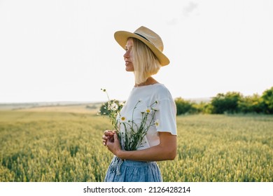Sensual blond caucasian woman in straw hat with a bouquet of chamomile enjoying summer in yellow field at sunset. Woman unity nature concept.