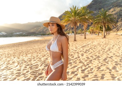 Sensual Brunette Woman In White Fashionable Bikini And Hat Posing At The Beach, Sunset Time. Summer Island Vibes. Travel.