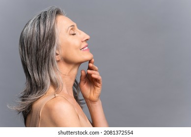 Sensual calm elderly woman stands in profile isolated on gray, smiling with eyes closed, touching chin with hand, gray-haired charming lady enjoying smooth of fresh skin after spa procedure