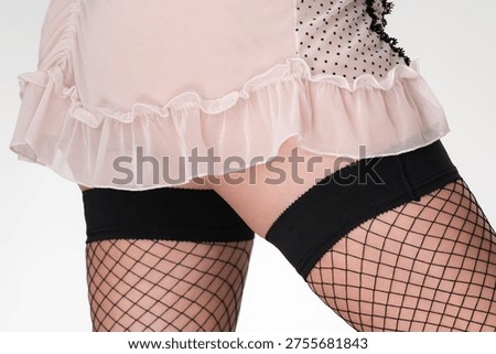 A sensual close-up view of a woman's upper thighs. She wears black fishnet stockings and a sheer, light pink babydoll with polka dots and ruffles. Studio shot on a white background. Evokes intimacy