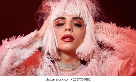 Sensual Drag Queen, Young Transgender With Rubbed Bright Night Makeup Wearing Boa Feather Party Dress And White Wig, Posing Isolated In Red Neon Light Background. Lgbtqi, Drag Show Concept