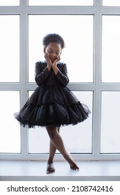 Sensual Little Female Kid Wearing Stylish Black Dress Standing On Windowsill With Bare Feet. African American Ballerina Holding Hands On Cheeks And Looking Down.