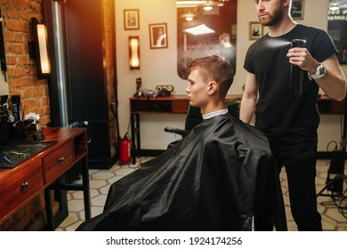 Sensual Manly Professional Hairdresser Spraying Client's Hair In A Barber Shop. Letting Drops Fall From Above. He's Standing Behind Client In A Chair Sitting In Profile.