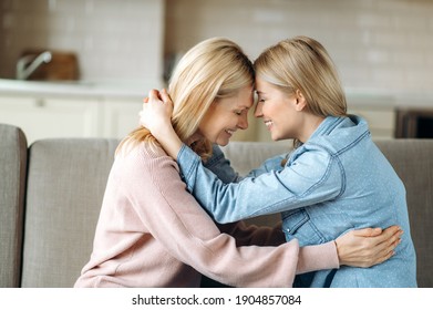 Sensual photo of aged blonde caucasian mother and her young adult happy daughter, are sitting at home on the couch at living room, embracing, smiling. Love of mom and daughter