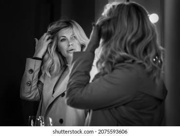 Sensual Portrait Of Middle Aged Seductive Woman In Trench Coat Gazing At Mirror Reflection Appreciating Her Hair Style. Human Emotion Or Clothing Fashion Concept Image. Back View Black And White Shot.