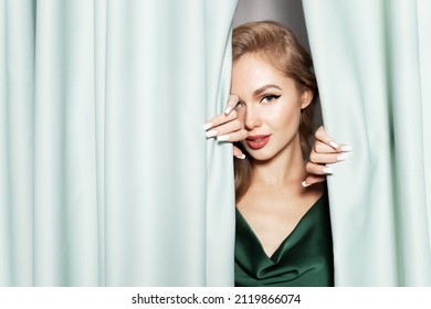 Sensual Pretty Young Blond Girl Wearing Celebratory Evening Dress, With Perfect Makeup Peek Out Green Curtains. Alluring Caucasian Female Posing Near Curtain. Festivity, Skincare Cosmetics Advertising