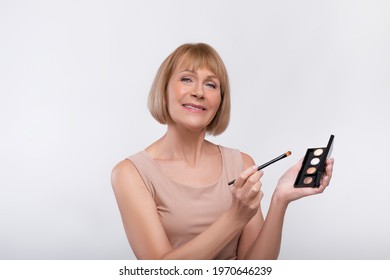 Sensual Senior Woman Holding Swatches Palette, Applying Eye Shadow With Brush On Light Studio Background. Beautiful Mature Lady Using Decorative Makeup, Recommending Cosmetic Product