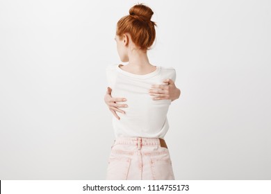 Sensual shot of attractive thin caucasian woman with natural red hair standing backwards to camera, hugging herself and turning head left, being upset with something, standing insecure over grey wall