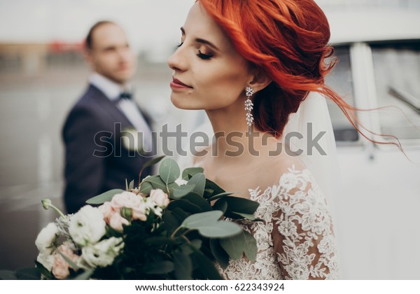 sensual stylish bride holding modern bouquet and\
relaxing with windy hair and groom looking at her near retro car.\
luxury wedding couple newlyweds posing. space for text. emotional\
moment