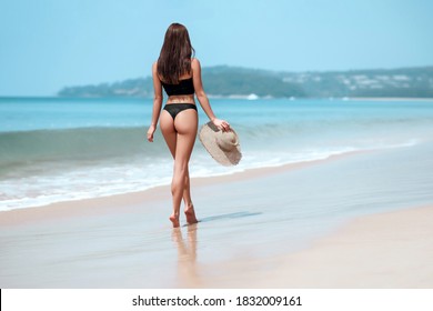 Sensual woman in black bikini walking along the coastline with straw hat in her hand