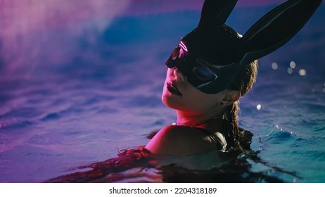 Sensual Woman In Leather BDSM Mask Moving Seductively In Swimming Pool Water Under Neon Color Light. Halloween Party, Attractive Chick Masquerade Bunny Enjoying Nighttime Games For Adults. High