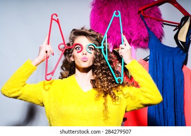 Sensual Young Girl With Curly Hair In Yellow Sweater Holding Hangers Standing Amid Colorful Clothes Pink Red Blue Colors On Grey Wall Background, Horizontal Picture