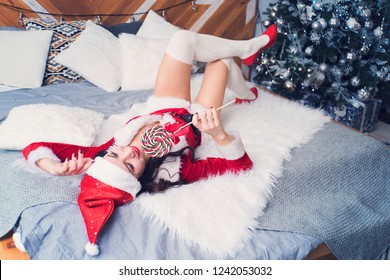Sensual young woman biting a candy cane. The girl in the red suit Santa is looking dreamy lying on the bed and takes in his mouth a Lollipop on a stick on background a decorated Christmas tree.