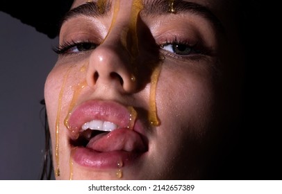 Sensual young woman face with honey drop close up. Sexy model with honey drip on face. Beauty woman face portrait in studio. Beautiful girl tongue licking honey lips.