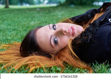 Sensual Young Woman With Gray Eyes Wearing A Silver Nose Ring Lying On The Green Lawn Of A Park, Spreading Out Her Long Hair With An Ombre Color Mix From Dark To Fiery Orange.