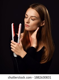 Sensual young woman with healthy silky straight hair stands with eyes closed holding white modern hair straightener in hand over dark background. Haircare, beauty, wellness concept