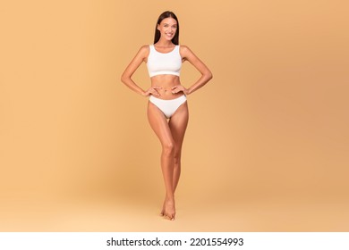 Sensual Young Woman Model Posing In White Underwear Over Beige Studio Background, Showing Perfect Figure And Smiling At Camera, Full Body Length, Copy Space