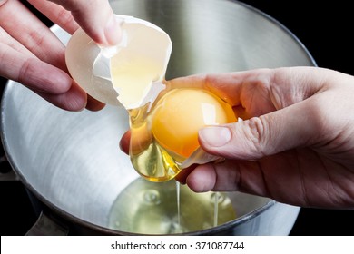 Separating Yolk And White Of The White Shell Egg With Woman Hands Above Metal Stainless Steel Mixing Bowl On Black Background