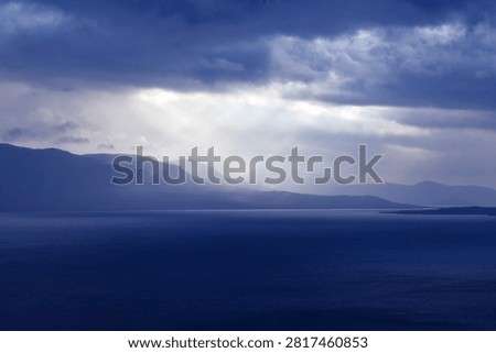 September, and as evening approaches, a few rays of sunshine break through the dark blue, rain-laden clouds. A lake lies on this side of the mountains in the background. 