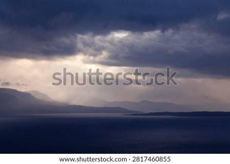 September, and as evening approaches, a few rays of sunshine break through the dark blue, rain-laden clouds. A lake lies on this side of the mountains in the background. 