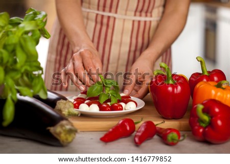 Ser mozzarella, pomidory na talerzu, kobieta w kuchni zdobi danie z zielonej bazylii. Obok znajdują się bakłażany, czerwona papryka chili i kolorowa papryka słodka.