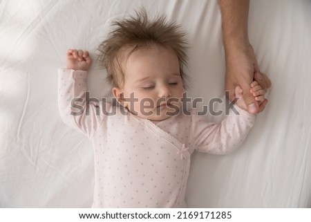 Serene adorable babygirl in pink bodysuit sleeping on bed holding tightly loving daddy thumb, touch finger feeling safety, close up above view. Family connection and ties, protection, cherish concept