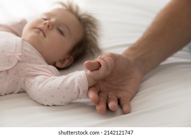 Serene adorable sleepy baby catching dads thumb, holding finger, lying on back, sleeping on bed. Cute infant child in pink bodysuit sleeping under parent supervision. Close up of hand, cropped shot