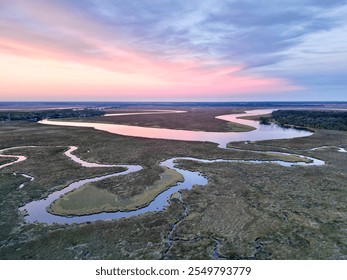 A serene aerial view of winding waterways through marshland at sunrise or sunset. Pastel pink and blue hues fill the sky, reflecting gently in the water, highlighting nature’s tranquil beauty. - Powered by Shutterstock