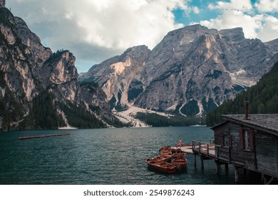 A serene alpine lake surrounded by rocky peaks and dense forests in the Italian Dolomites. Wooden boats rest at a dock beside a rustic cabin, creating a tranquil and picturesque mountain retreat. - Powered by Shutterstock