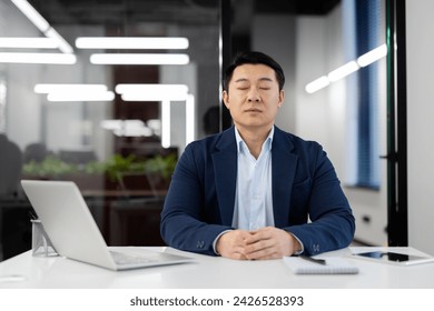 Serene Asian businessman practicing mindfulness meditation in modern office setting to alleviate stress and maintain focus. - Powered by Shutterstock