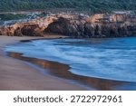 A serene and atmospheric view of the dramatic cliffs surrounding Praia Pintadinho in the Algarve, Portugal, where the golden glow of sunset or sunrise bathes the layered rock formations in warm hues. The rugged coastline features a series of natural sea caves and arches carved by centuries of erosion, standing majestically above the calm, turquoise waters. Gentle waves wash over the sandy beach below, creating a soft, blurred motion that contrasts with the solid, ancient stone. Lush green vegetation crowns the cliff top, completing this tranquil coastal scene — ideal for travel, nature, or cinematic backgrounds.