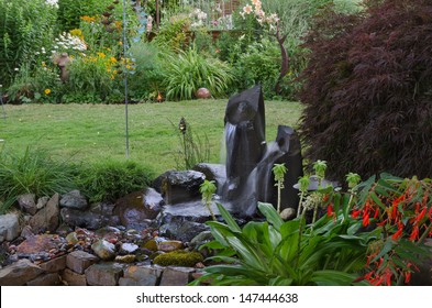 A Serene Backyard Retreat With Twin Fountains In The Foreground And A County Garden In The Background With Several Pieces Of Art Nestled Among The Flowers. 