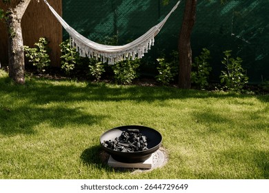 A serene backyard setting features a white macrame hammock strung between trees, a black fire pit, and a vibrant green lawn, inviting outdoor relaxation and enjoyment