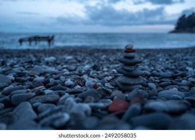 A serene beach scene at dusk, featuring a carefully balanced stack of stones on a pebble-strewn shore. The moody blue sky and calm sea create a tranquil backdrop, with a distant pier silhouette. - Powered by Shutterstock