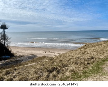 A serene beach scene with gentle waves approaching the sandy shore, grassy dunes in the foreground, and a vast horizon under a partly cloudy sky. - Powered by Shutterstock