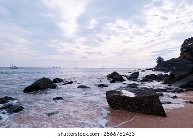 A serene beach scene with rocks and gentle waves under a cloudy sky, with sailboats in the distance. - Powered by Shutterstock