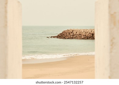 A serene beach scene with a sandy shore and a breakwater made of large concrete blocks extending into the calm sea, framed by two white pillars. - Powered by Shutterstock