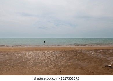 Serene beach scene: a solitary figure stands in calm waters as rippled sand and scattered debris frame the tranquil ocean beneath a partly cloudy sky. Bushehr, Iran.  - Powered by Shutterstock - Get 15% off with code: PIKWIZARD15