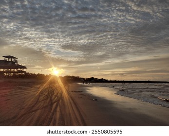 A serene beach scene at sunset with a charming wooden structure silhouetted against the glowing sky. The sun dips below the horizon, casting warm light and creating a breathtaking display of clouds. - Powered by Shutterstock