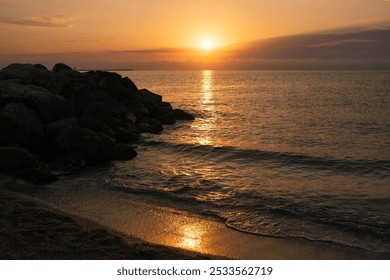 A serene beach scene at sunset with a rocky shoreline and calm waves under an orange sky. - Powered by Shutterstock