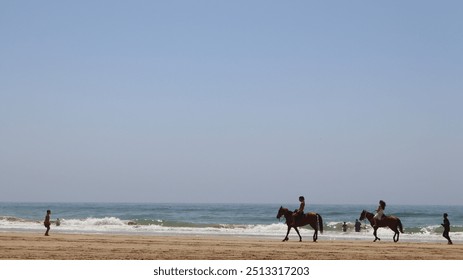 A serene beach scene with two horseback riders by the shore, gentle waves lapping the sand, a clear sky, and distant figures enjoying the water, evoking tranquility and the beauty of nature. - Powered by Shutterstock