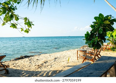 A serene beachside setting featuring two wooden lounge chairs positioned under lush tropical palm leaves. The chairs are set on a pristine sandy beach with gentle waves lapping at the shore. Clear blue skies and calm waters create a tranquil atmosphere, perfect for vacation advertisements, travel brochures, or wellness retreats. The natural shade from the palm leaves enhances comfort, making it ideal for relaxation or beach-themed promotions.