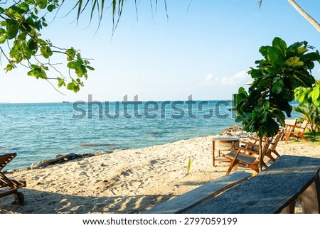 A serene beachside setting featuring two wooden lounge chairs positioned under lush tropical palm leaves. The chairs are set on a pristine sandy beach with gentle waves lapping at the shore. Clear blue skies and calm waters create a tranquil atmosphere, perfect for vacation advertisements, travel brochures, or wellness retreats. The natural shade from the palm leaves enhances comfort, making it ideal for relaxation or beach-themed promotions.