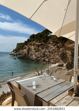 A serene beachside table set for two at an Ibiza beach club, overlooking a rocky cove and crystal-clear water, shaded by a white umbrella. Perfect for a tranquil seaside dining experience.